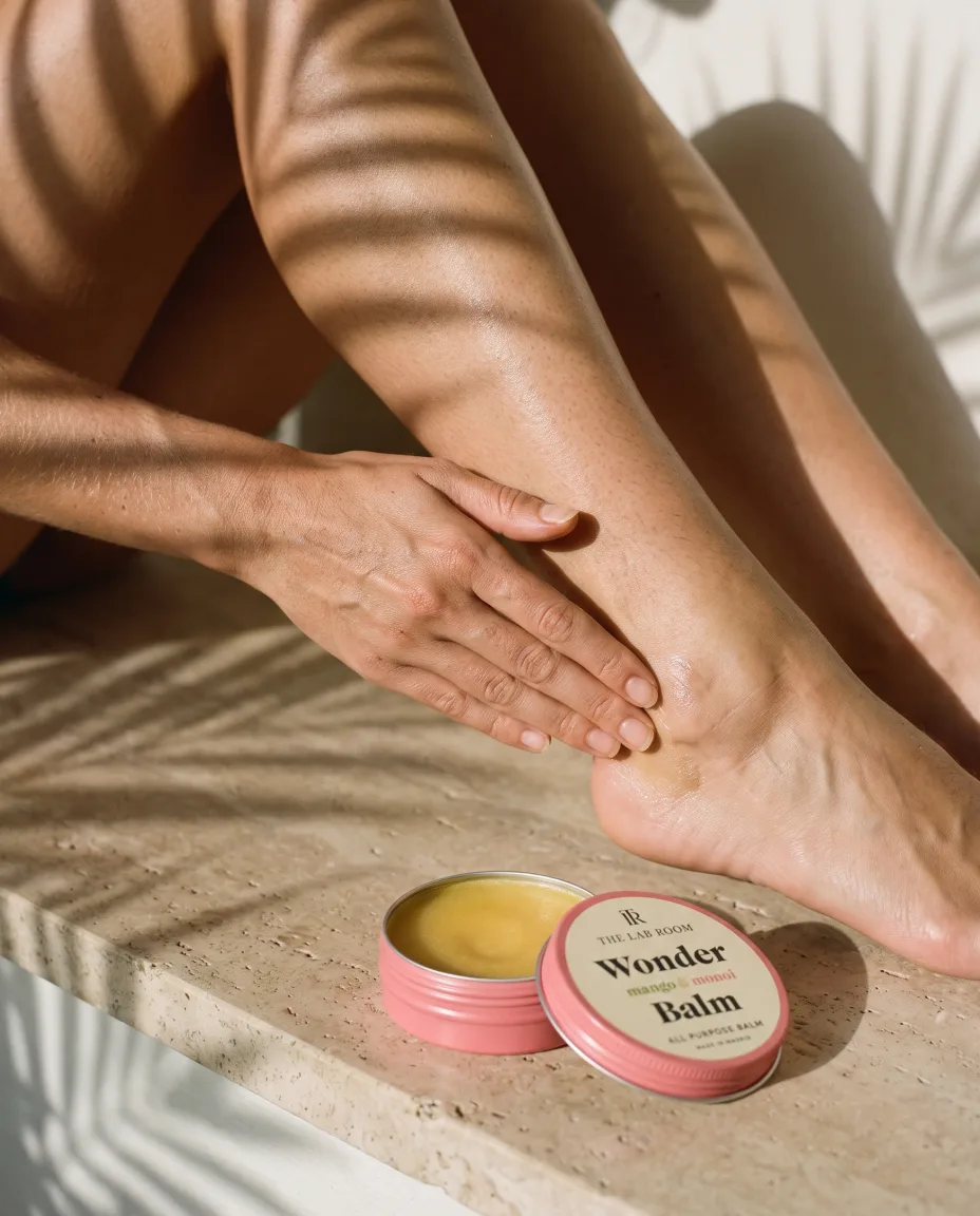 Hand smoothing the balm over the ankle and heel, with the tin on travertine